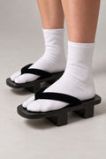 All Black Geta Footwear 'Shikkokuba' – all black wooden geta with plain black strap, two-tooth wooden geta sandals with padded fabric strap, traditional Japanese footwear, worn with white tabi sock, side view on model
