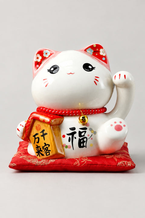 Beckoning Cat Money Box 'Manekite' – round-eyed white ceramic lucky cat money box with one paw beckoning and a gold sign, Japanese maneki neko lucky cat figurine on a red brocade cushion, studio view