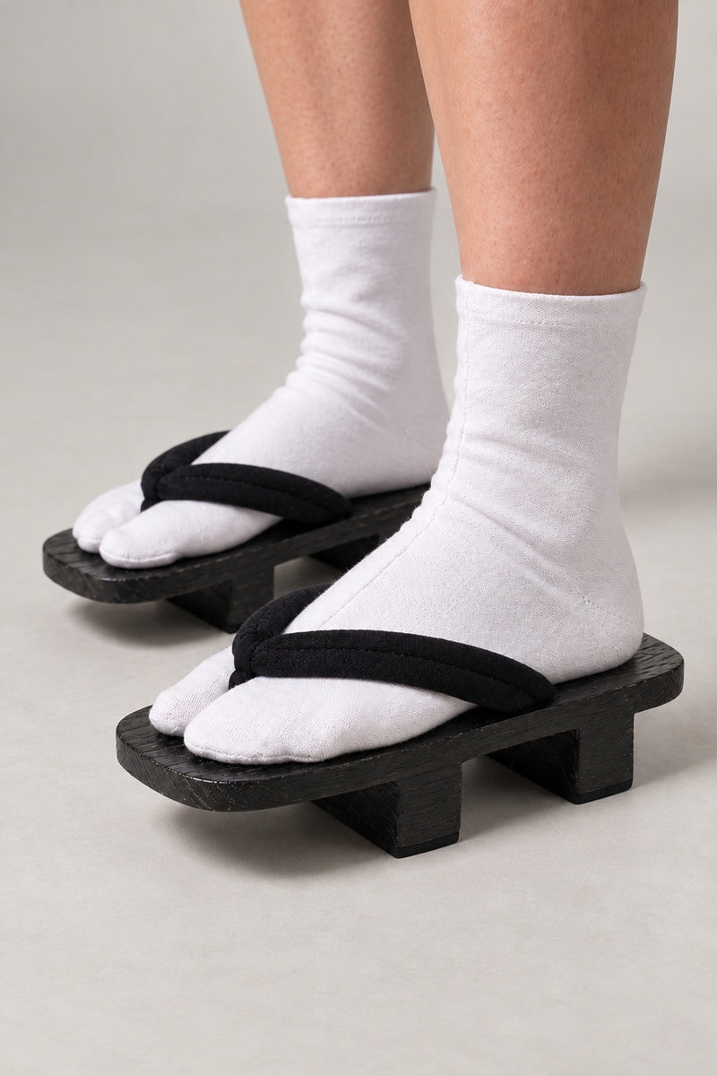 Black Corded Traditional Geta 'Kuronawa' – black wooden geta with black corded strap, plain traditional two-tooth wooden geta sandals with padded fabric strap, traditional Japanese footwear, worn with white tabi sock, side view on model