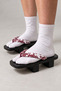Black Geta Sandals 'Benibana' – black wooden geta with white strap printed in red floral scroll, two-tooth wooden geta sandals with padded fabric strap, traditional Japanese footwear, worn with white tabi sock, side view on model