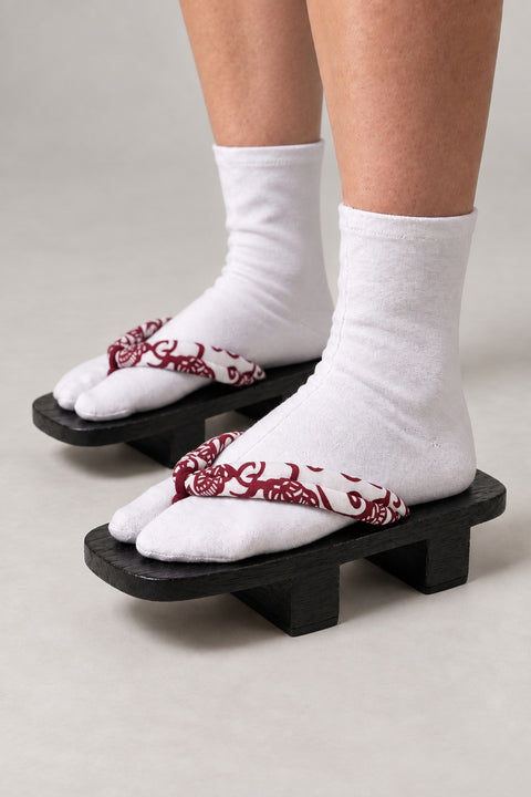 Black Geta Sandals 'Benibana' – black wooden geta with white strap printed in red floral scroll, two-tooth wooden geta sandals with padded fabric strap, traditional Japanese footwear, worn with white tabi sock, side view on model