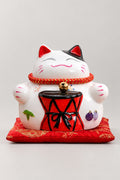 Festival Drum Maneki Neko 'Taikoneko' – calico ceramic lucky cat holding a red festival drum in both paws, Japanese maneki neko lucky cat figurine on a red brocade cushion, studio view