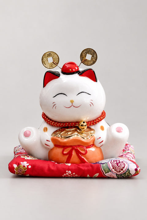Fortune Bag Maneki Neko 'Kinchaku' – white ceramic lucky cat holding an orange fortune bag with nodding gold coins above its head, Japanese maneki neko lucky cat figurine on a red brocade cushion, studio view