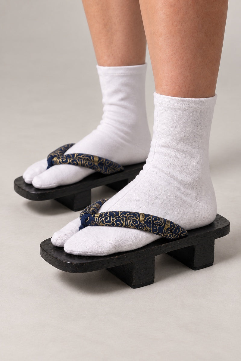 Gold Karakusa Geta Japanese Shoes 'Kinkusa' – black wooden geta with navy strap printed in gold karakusa vine scroll, two-tooth wooden geta sandals with padded fabric strap, traditional Japanese footwear, worn with white tabi sock, side view on model