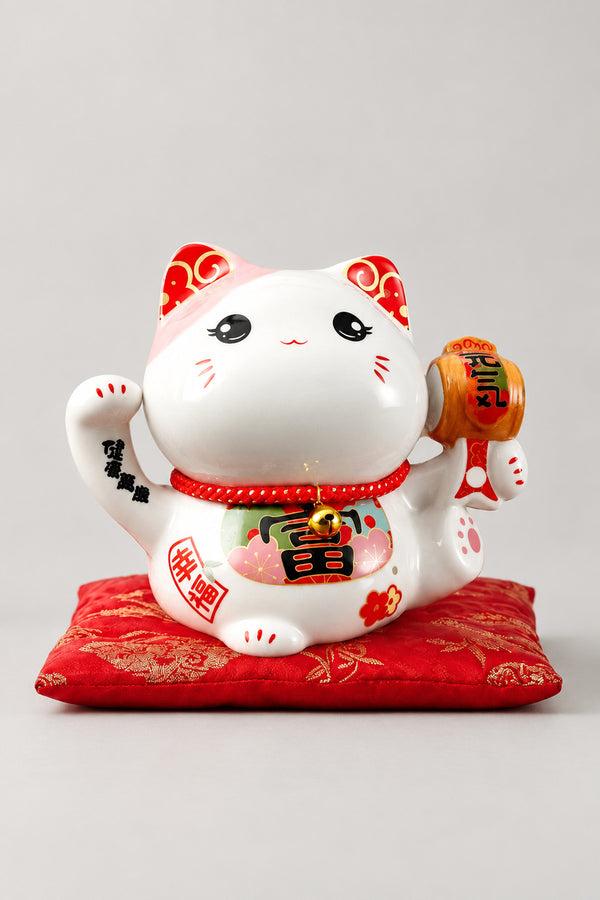 Golden Mallet Lucky Cat Bank 'Kozuchi' – round-eyed white ceramic lucky cat bank holding a gold mallet with a red fortune character on the belly, Japanese maneki neko lucky cat figurine on a red brocade cushion, studio view