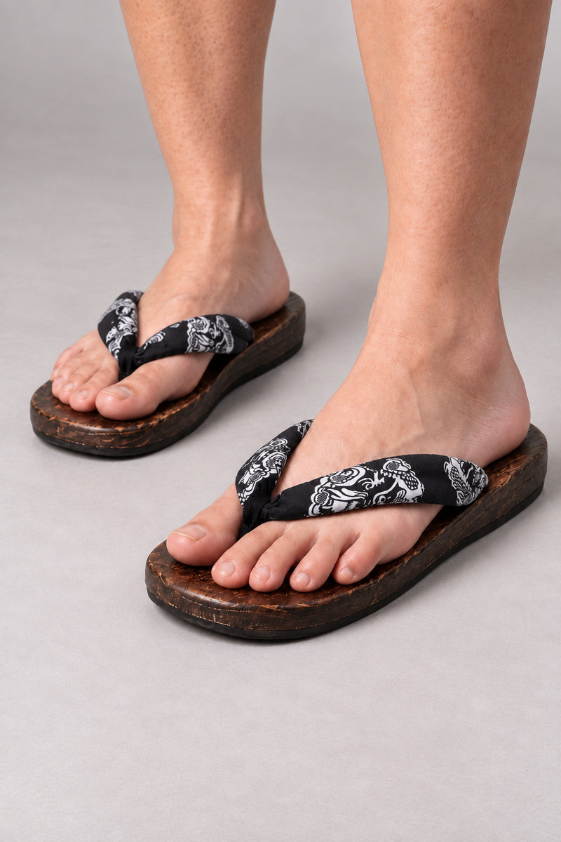 Japanese Flip Flops 'Karakoro' – wooden flip flops with black paisley print thong, flat Japanese wooden sandals worn barefoot, front view on model