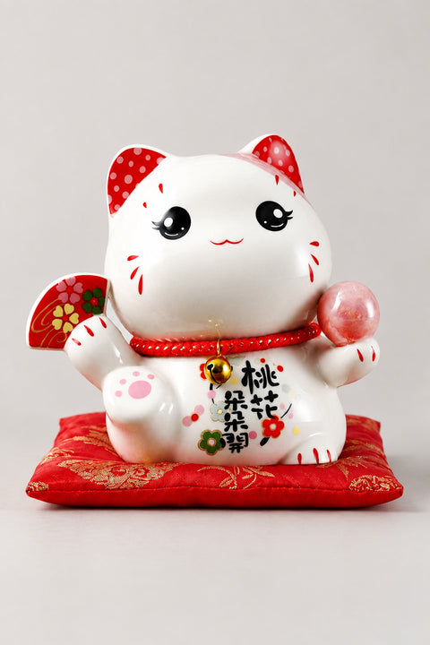 Kawaii Maneki Neko Piggy Bank 'Tamaneko' – round-eyed white ceramic lucky cat piggy bank holding a red fan and pink ball, Japanese maneki neko lucky cat figurine on a red brocade cushion, studio view