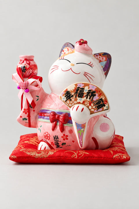 Large Pink Maneki Neko Statue 'Ooneko' – large ceramic lucky cat statue in a painted pink kimono with red bow holding a fan and bell charm, Japanese maneki neko lucky cat figurine on a red brocade cushion, studio view