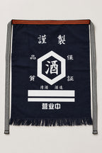 Maekake – indigo maekake apron printed with a round sake crest and quality seal, traditional Japanese sake brewery waist apron with fringed hem, studio view