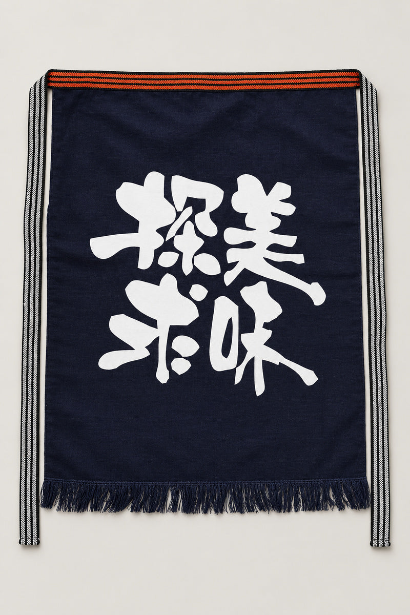 Maekake – indigo maekake apron printed with two bold brushed kanji for delicious, traditional Japanese sake brewery waist apron with fringed hem, studio view