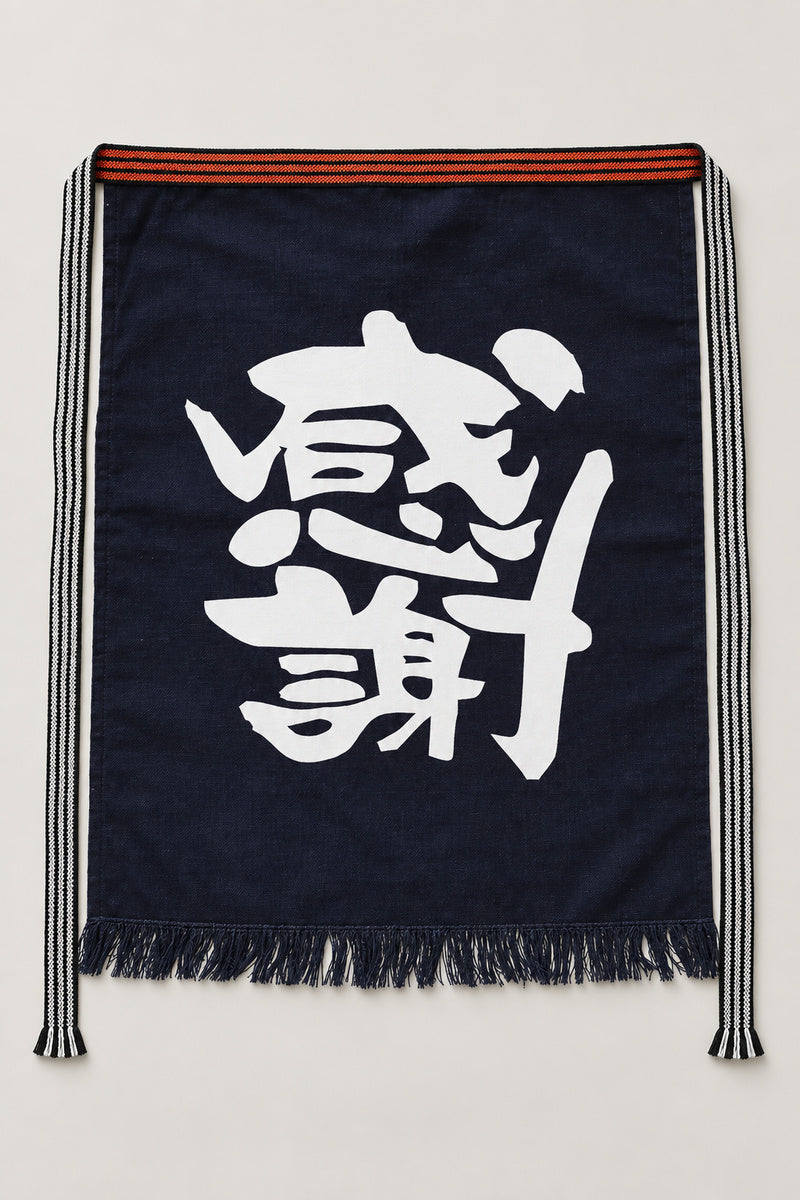 Maekake – indigo maekake apron printed with the bold kanji for gratitude, traditional Japanese sake brewery waist apron with fringed hem, studio view