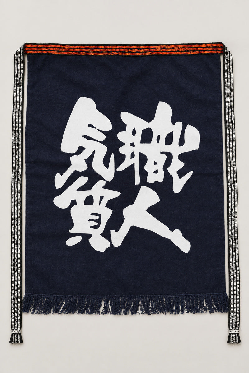 Maekake – indigo maekake apron printed with the kanji for craftsman spirit, traditional Japanese sake brewery waist apron with fringed hem, studio view