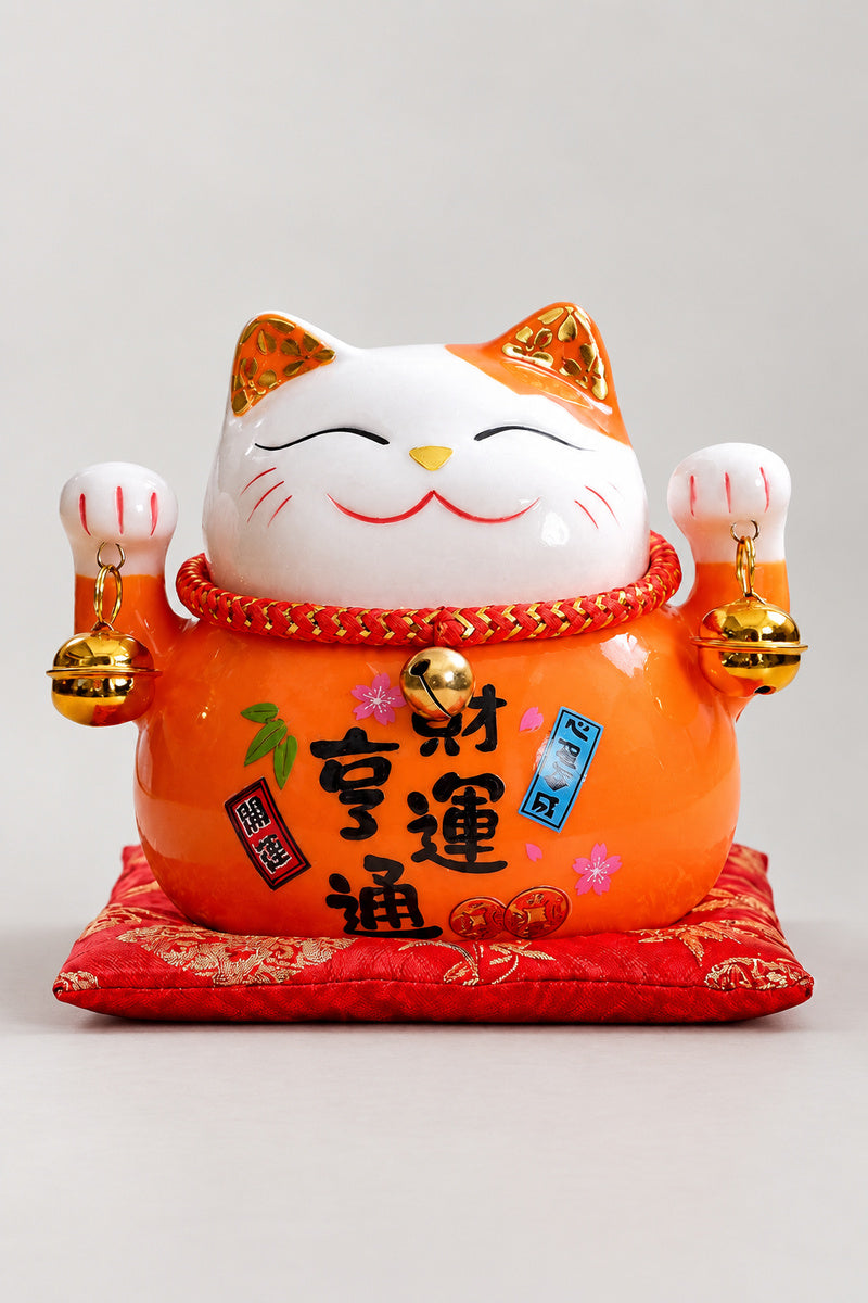 Orange Maneki Neko Figurine 'Daidaineko' – orange ceramic lucky cat with both paws raised and a brushed wish for money luck, Japanese maneki neko lucky cat figurine on a red brocade cushion, studio view