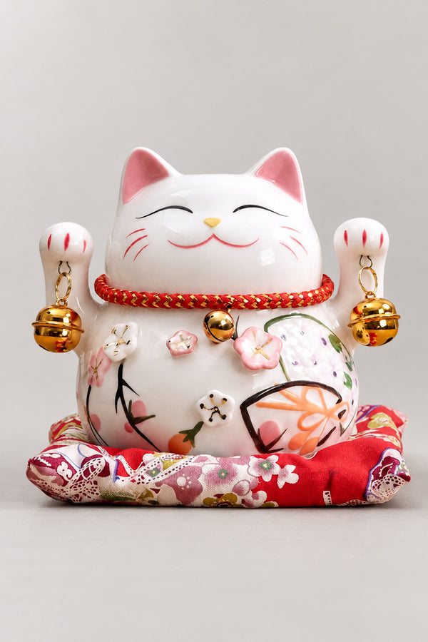 Plum Blossom Maneki Neko 'Umeneko' – white ceramic lucky cat with both paws raised, plum blossom and fan painted on the body, red collar and brass bells, Japanese maneki neko lucky cat figurine on a red brocade cushion, studio view