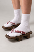 Red Strap Geta Sandals 'Akabana' – brown wooden geta with white strap printed in red floral scroll, two-tooth wooden geta sandals with padded fabric strap, traditional Japanese footwear, worn with white tabi sock, side view on model