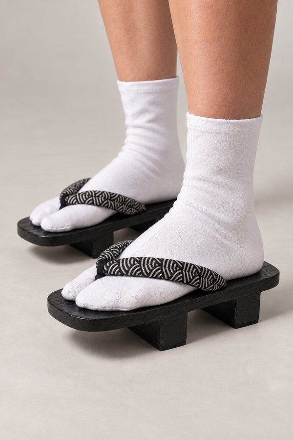 Seigaiha Japanese Footwear Geta 'Kuronami' – black wooden geta with black strap printed in grey seigaiha wave pattern, two-tooth wooden geta sandals with padded fabric strap, traditional Japanese footwear, worn with white tabi sock, side view on model