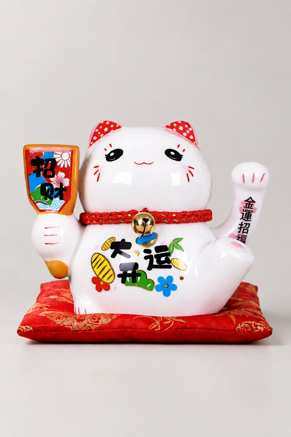 Solar Waving Maneki Neko 'Teoneko' – white solar powered waving lucky cat holding a sign with a wish painted on the body, Japanese maneki neko lucky cat figurine on a red brocade cushion, studio view